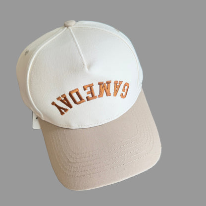 Two caps, one cream and one dark beige, with 'GAMEDAY' text on a baseball hat. 